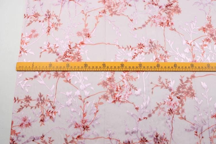 a close-up view of a fabric with a floral pattern, featuring pink and red flowers and leaves. A yellow measuring tape is laid across the fabric, indicating the size or dimensions of the fabric.