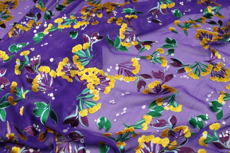 a close-up view of a fabric with a vibrant floral pattern in shades of purple, yellow, and green. The pattern is composed of numerous small flowers and leaves, creating a lively and colorful visual effect. The fabric appears to be made of silk or a similar material, and the intricate design suggests a high level of craftsmanship.