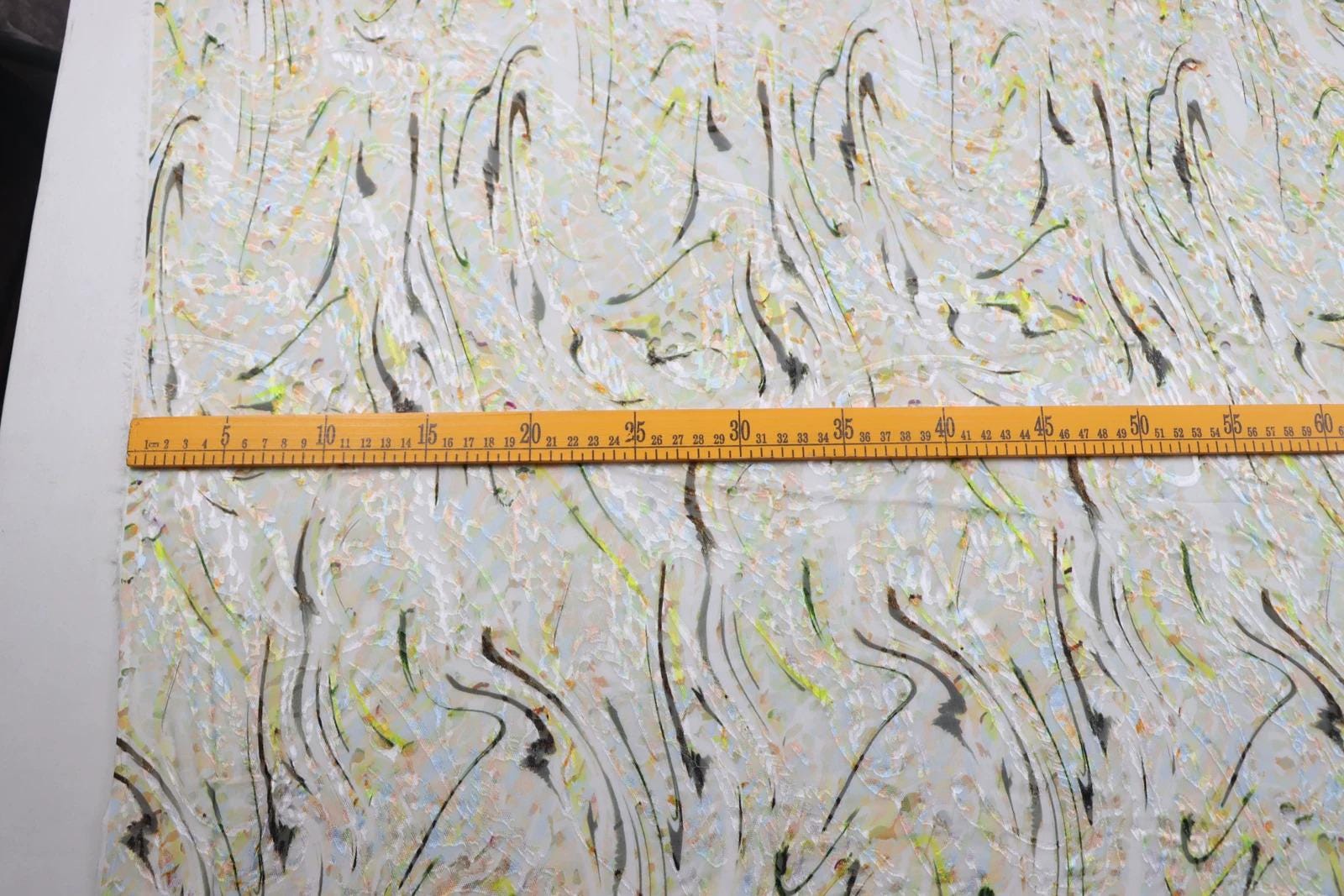 a close-up view of a fabric with a colorful, abstract pattern. A yellow measuring tape is placed across the fabric, likely for size or scale purposes.