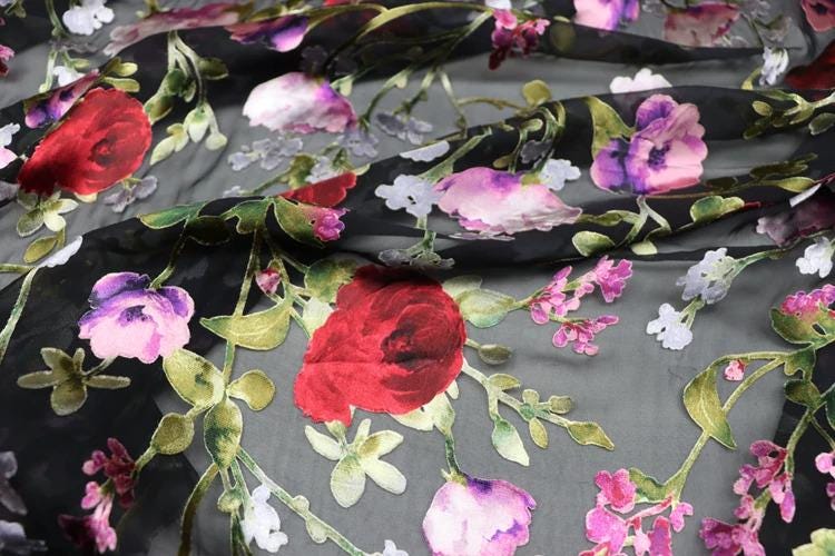 a close-up view of a fabric with a floral pattern, including red and pink flowers, green leaves, and white flowers. The fabric appears to be black, with the flowers and leaves in shades of red, pink, and white. The image does not contain any text or other discernible objects or actions.