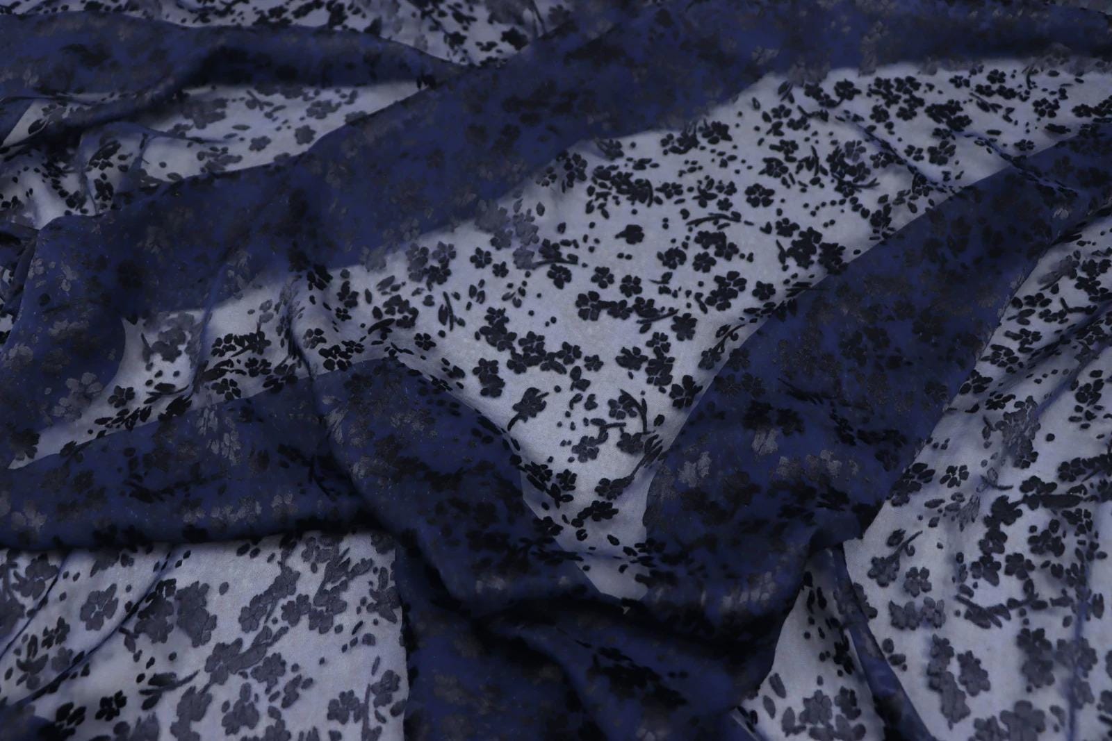 a close-up view of a fabric with a floral pattern in shades of blue and white. The fabric appears to be made of sheer material, with a dark blue color dominating the background. The pattern consists of small black flowers or leaves scattered across the fabric, creating a visually striking contrast against the lighter background.