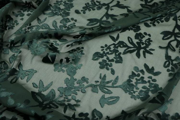 a close-up view of a fabric with a floral pattern, featuring green leaves and flowers. The fabric appears to be made of a material with a green color, possibly a type of lace or a similar textile. The pattern is intricate and detailed, with the leaves and flowers covering the entire fabric surface.