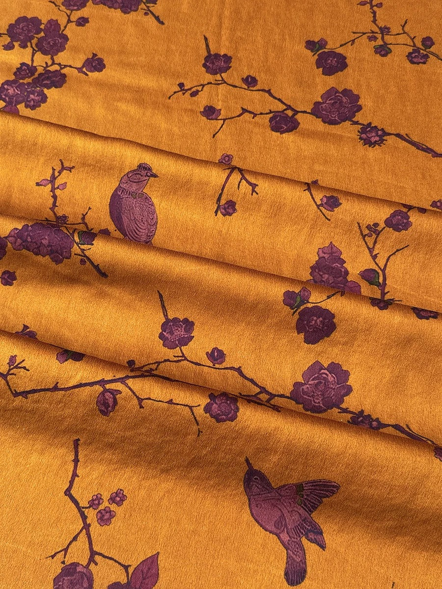 a close-up view of a fabric with a repeating pattern of purple birds perched on branches with flowers. The birds appear to be in various poses, creating a dynamic and visually interesting design. The fabric has a warm, golden-brown color that contrasts with the purple hues of the birds and flowers, making the pattern stand out.