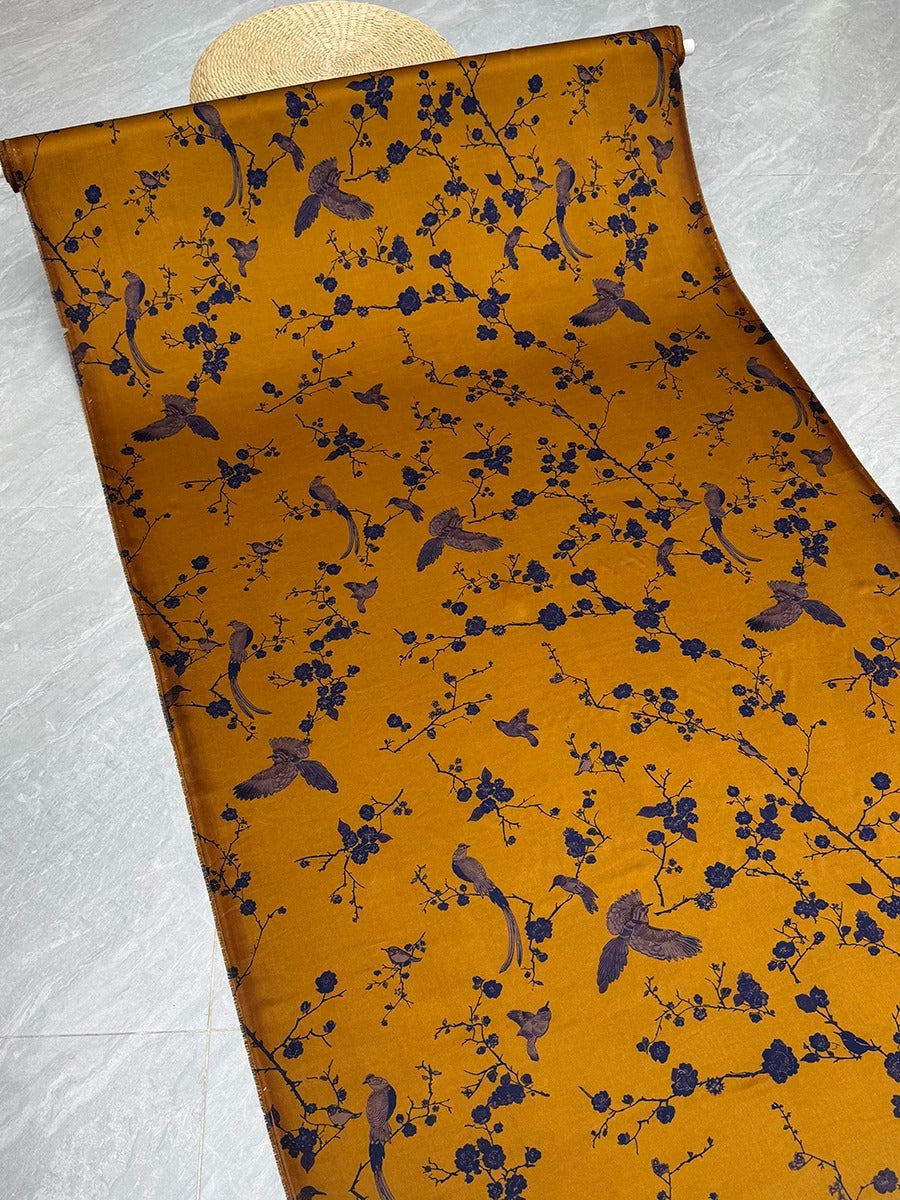 a vibrant yellow fabric with a repeating pattern of blue birds and flowers. The fabric appears to be rolled up and displayed on a white surface.
