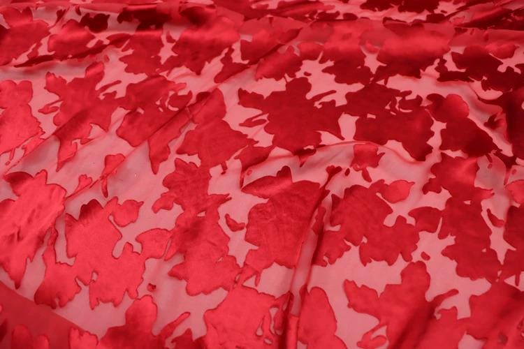 a close-up view of a fabric with a red floral pattern. The fabric appears to be made of a shiny, satin-like material, and the pattern consists of red flowers or leaves against a pink background. The fabric is crumpled, giving it a textured appearance.
