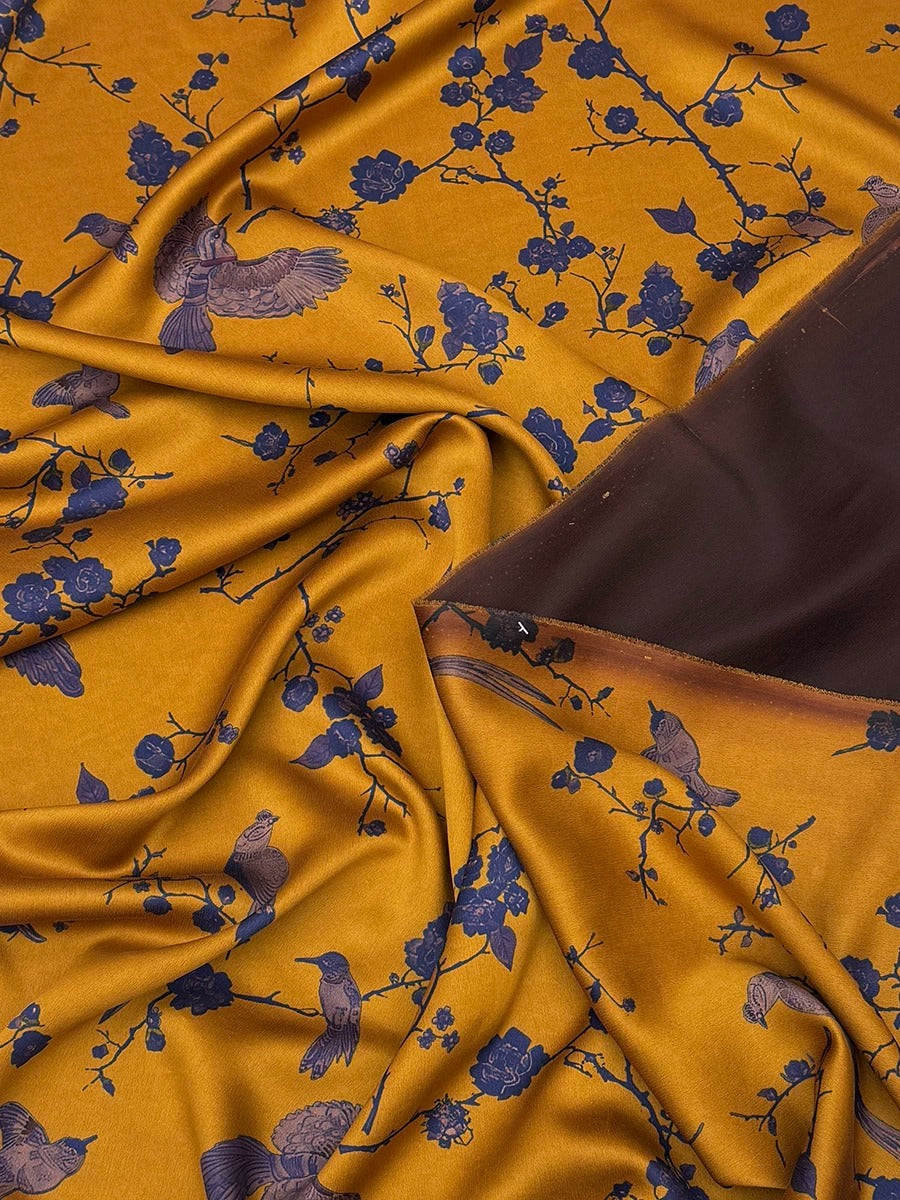 a close-up view of a fabric with a floral pattern in shades of yellow and blue. The fabric appears to be made of silk or a similar material, and it has a crinkled texture. The pattern consists of blue flowers and branches, creating a visually striking contrast against the yellow background. The fabric is folded and crumpled, giving it a soft and delicate appearance.