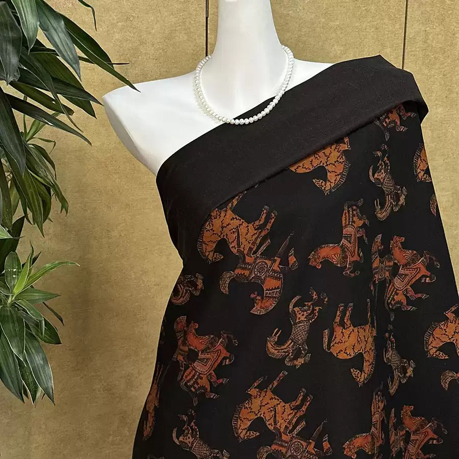 A mannequin wearing a black dress with a pattern of orange and brown animals, such as elephants and tigers, printed on it. The dress has a black top and a black skirt. The mannequin is positioned in front of a plant, creating a contrast between the mannequin and the natural elements.