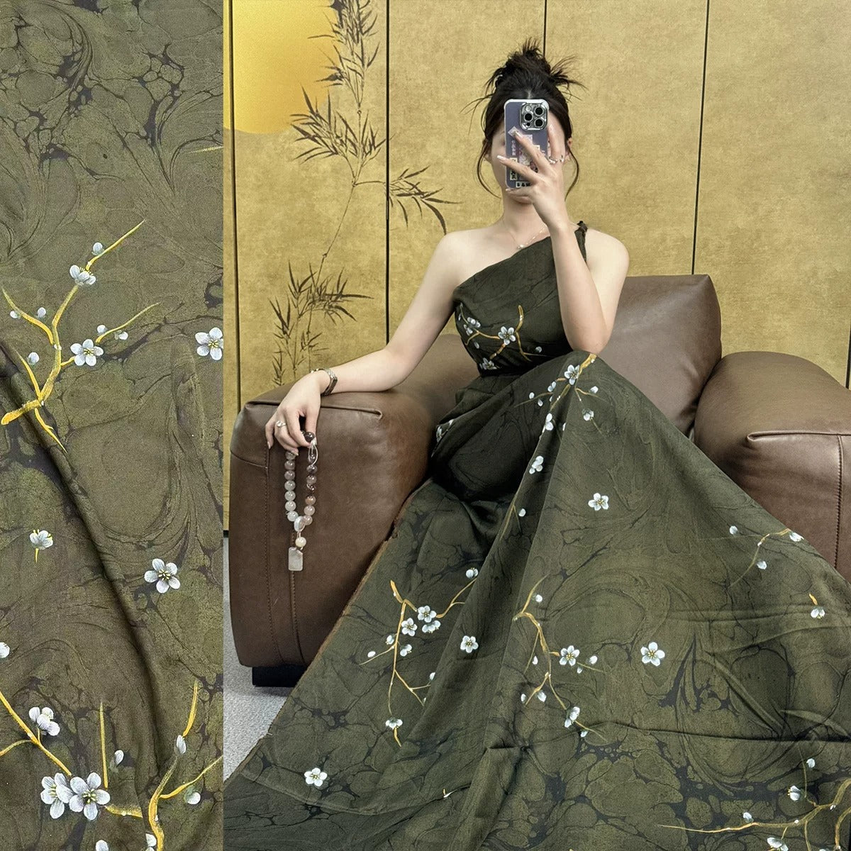 A woman in a green dress with white flowers is sitting on a brown leather couch, holding a cell phone and taking a selfie.