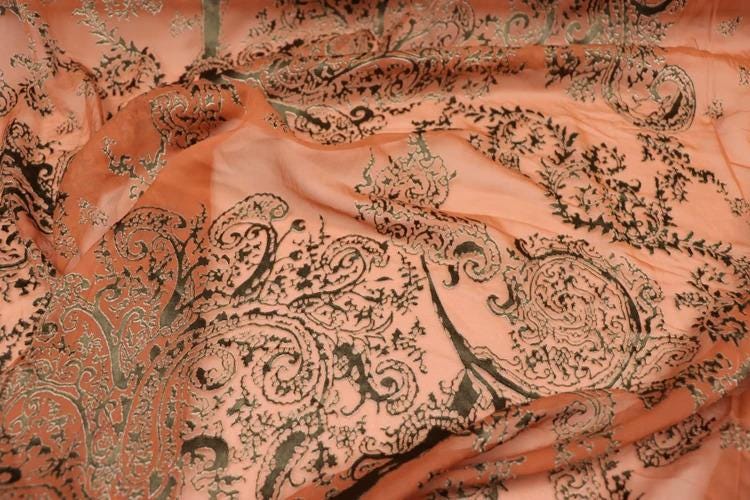 a close-up view of a fabric with a floral pattern in shades of orange and black. The fabric appears to be made of silk or a similar material, and the intricate design features a repeating pattern of leaves and flowers. The image does not contain any text or other discernible objects or actions.
