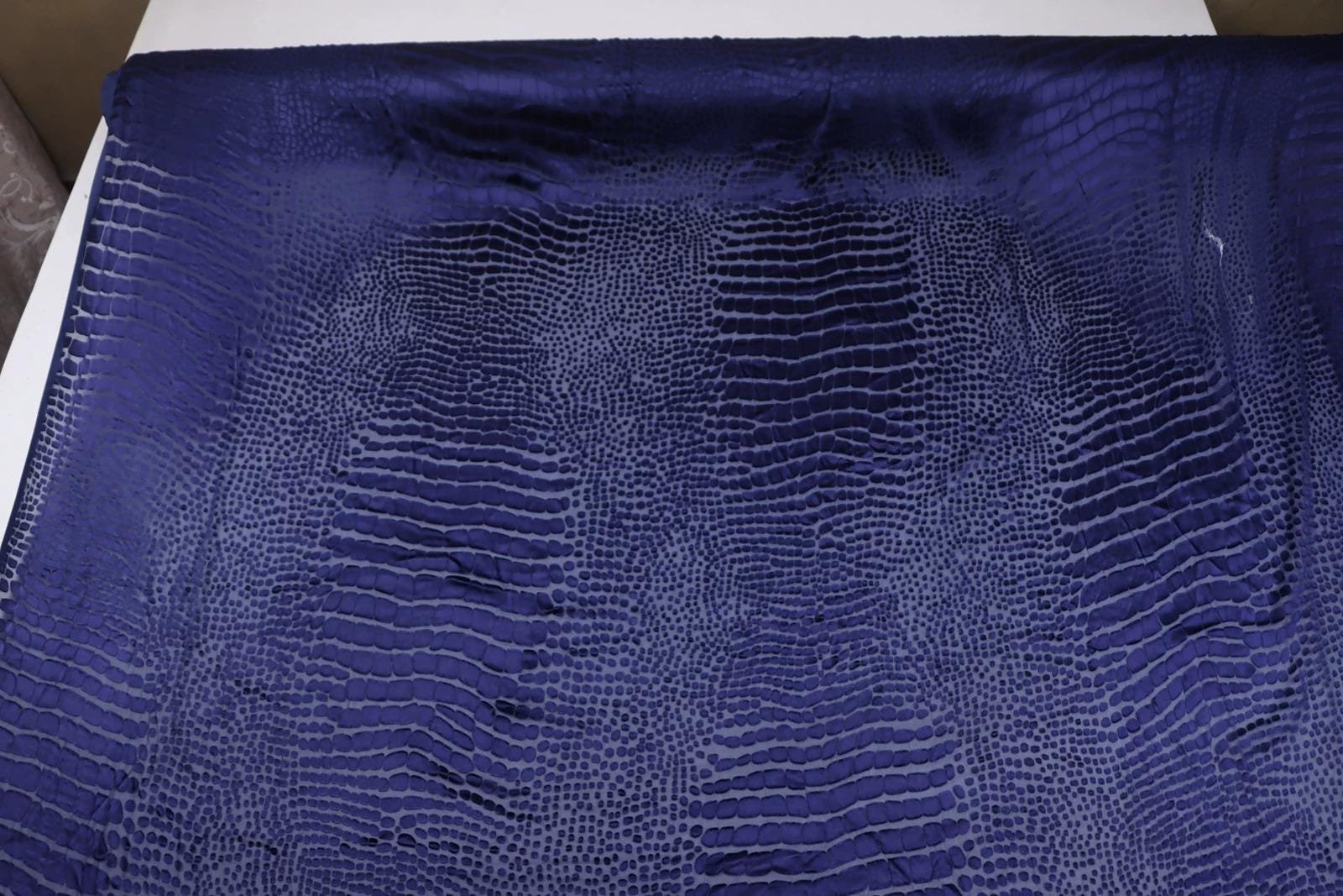 a close-up view of a fabric with a textured, patterned design in shades of blue. The fabric appears to be a type of leather or a similar material, with a distinctive pattern that resembles scales or skin texture.