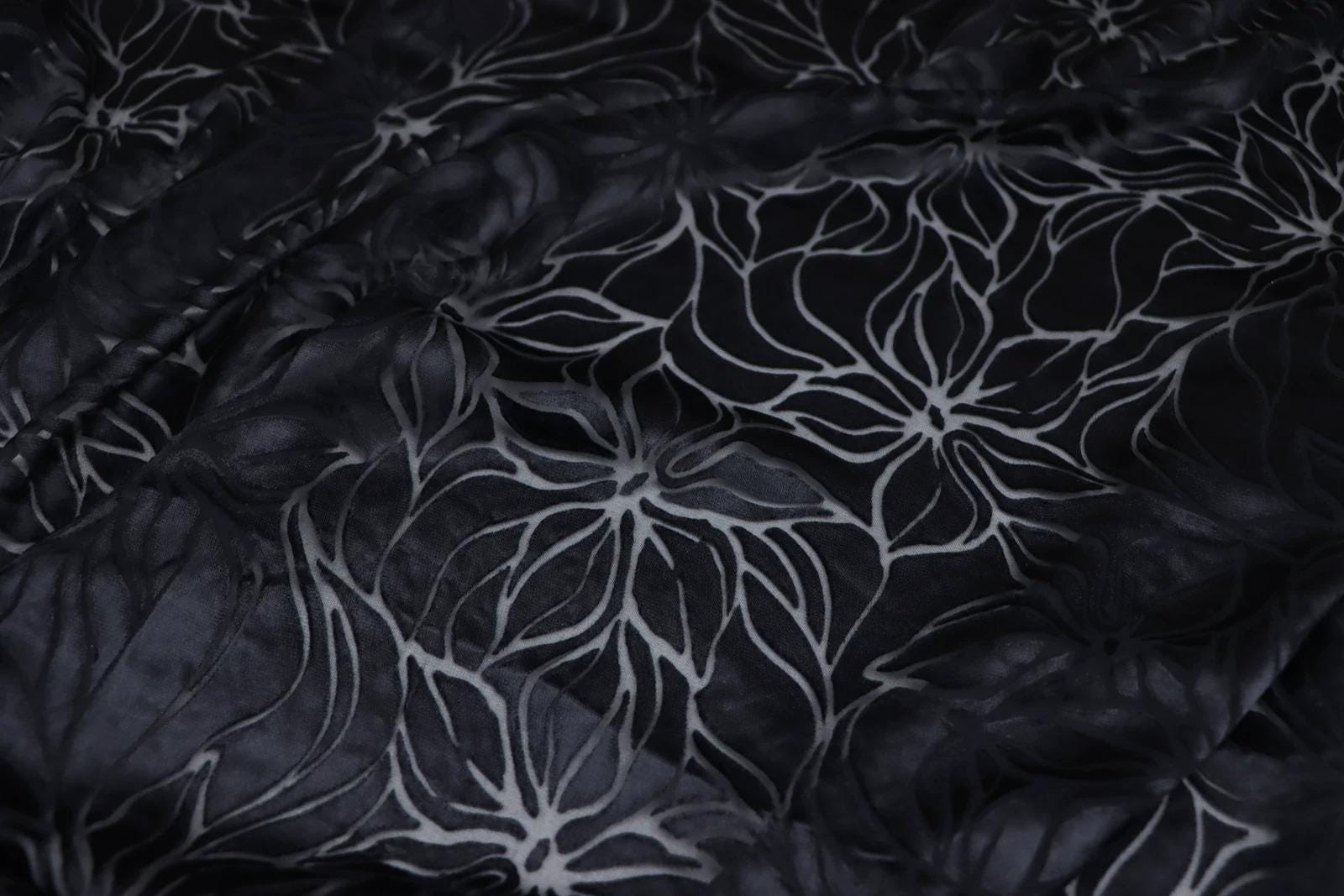 a close-up view of a fabric with a black and white floral pattern. The fabric appears to be made of a shiny material, possibly silk or satin, and has a textured appearance. The pattern consists of repeating floral motifs, creating a visually striking and intricate design.