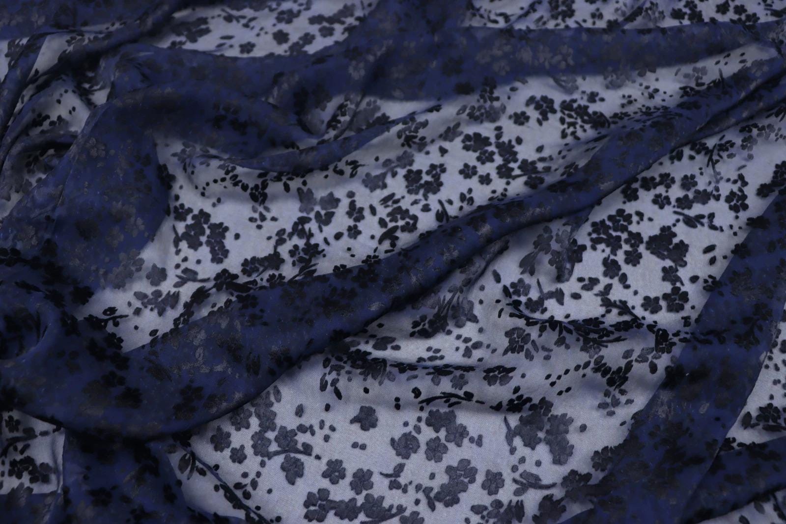 a close-up view of a fabric with a floral pattern in shades of blue and white. The fabric appears to be made of silk or a similar material, and the pattern consists of small black flowers or leaves against a white background. The fabric is crumpled and wrinkled, giving it a textured and soft appearance.