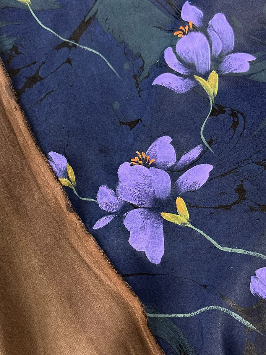 a close-up view of a fabric with a striking purple flower design. The fabric has a dark blue background with a brownish-brown border, creating a visually appealing contrast. The purple flowers are the main focus of the image, with their vibrant color and intricate details clearly visible.