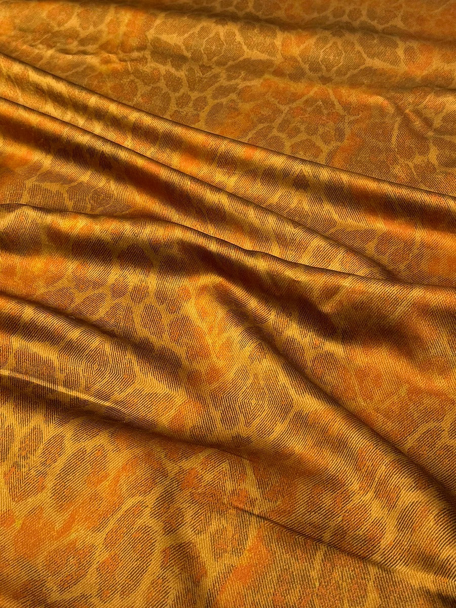 a close-up view of a fabric with a distinctive orange and brown pattern, resembling the design of a leopard or a similar animal. The fabric appears to be made of silk or a similar material, and it has a crinkled texture, giving it a soft and luxurious appearance.