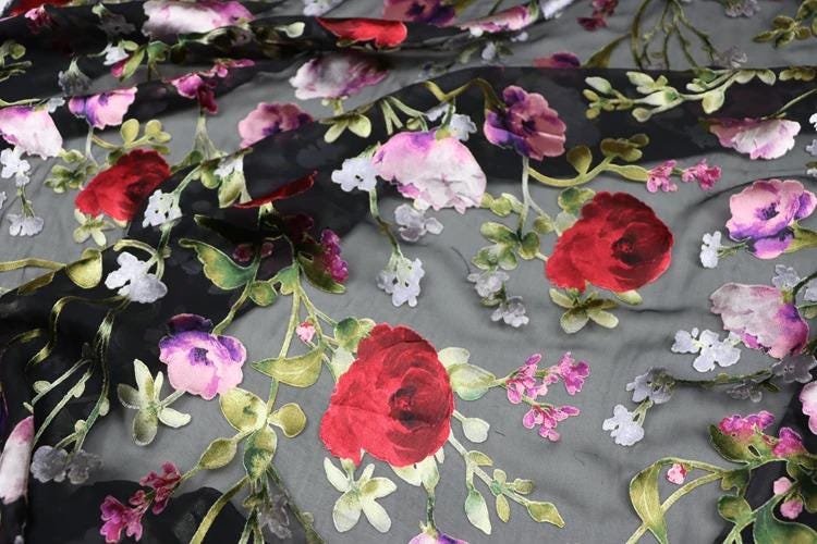 a close-up view of a fabric with a floral pattern. The fabric is predominantly black, with red and white flowers and leaves scattered across it. The flowers appear to be the main focus of the image, with the leaves providing a contrasting background. The overall composition of the image is visually appealing and showcases the intricate details of the floral pattern.
