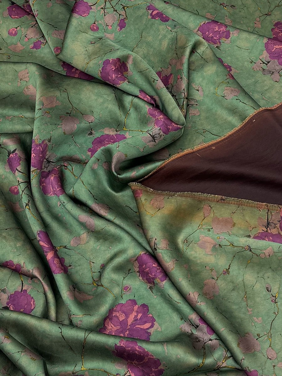 a close-up view of a fabric with a floral pattern in shades of green and purple. The fabric appears to be made of silk or a similar material, and it has a crinkled texture. The pattern consists of various shades of green and purple flowers, creating a visually appealing and intricate design.
