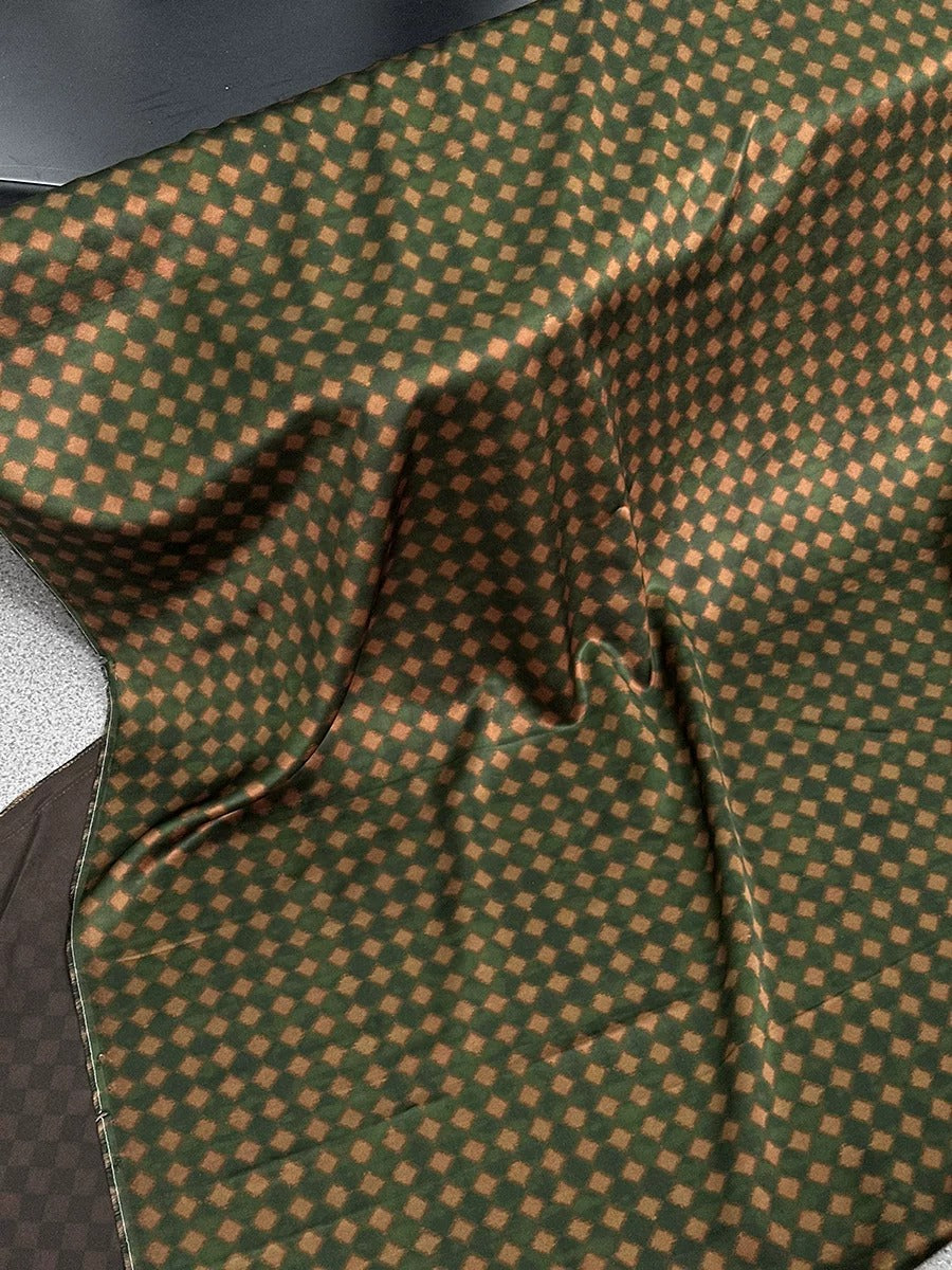 a close-up view of a fabric with a distinctive green and brown checkered pattern. The fabric appears to be made of a soft, textured material, possibly cotton or linen, and has a slightly wrinkled appearance. The pattern consists of small squares or diamonds arranged in a diagonal line across the fabric, creating a visually interesting and textured surface.