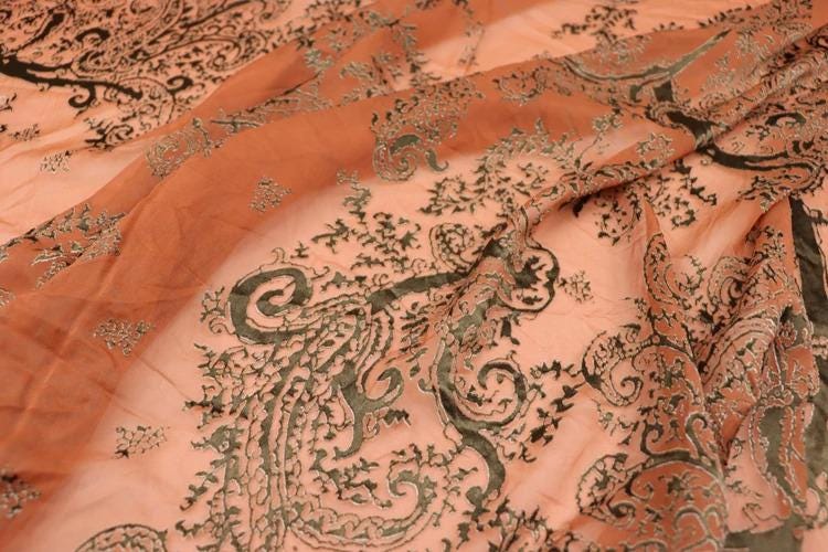 a close-up view of a fabric with a floral pattern in shades of orange and black. The fabric appears to be made of silk or a similar material, and the intricate design features a mix of swirling and curling patterns. The overall composition of the image is visually appealing and showcases the craftsmanship of the fabric.