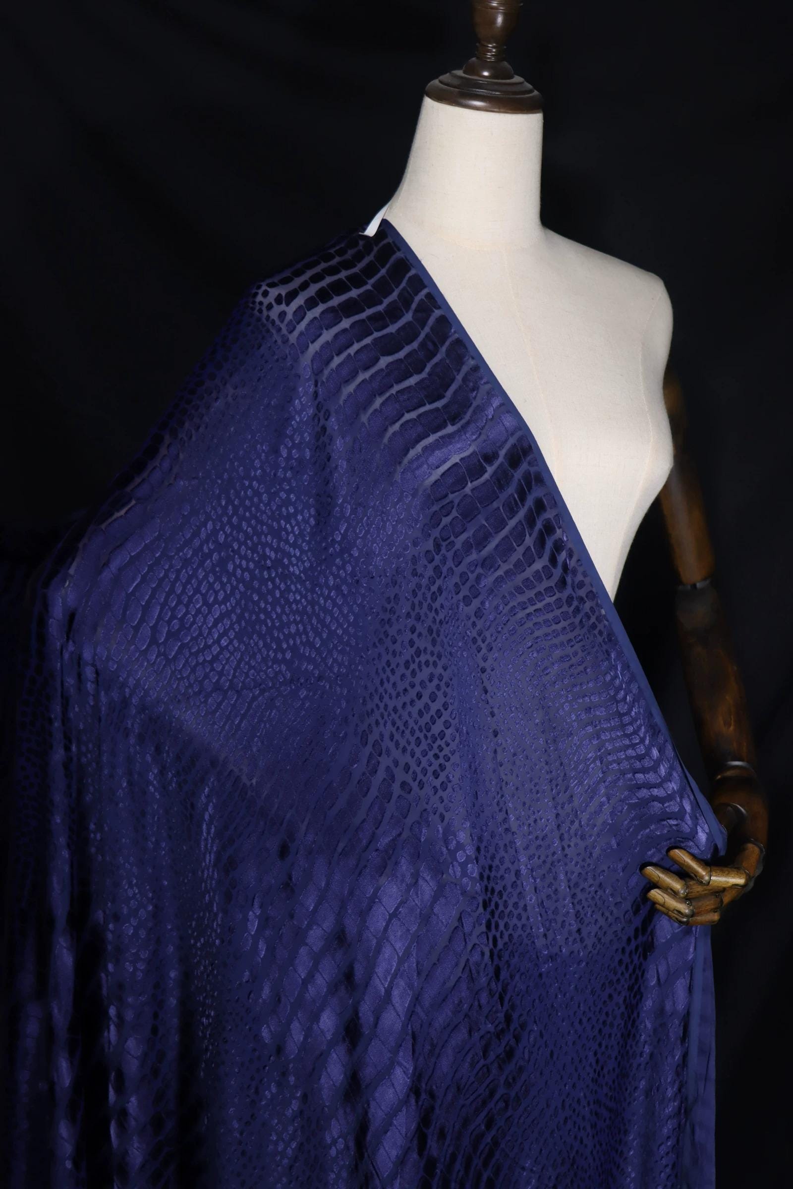 A mannequin wearing a blue fabric with a pattern resembling scales or snakes, displayed against a dark background.