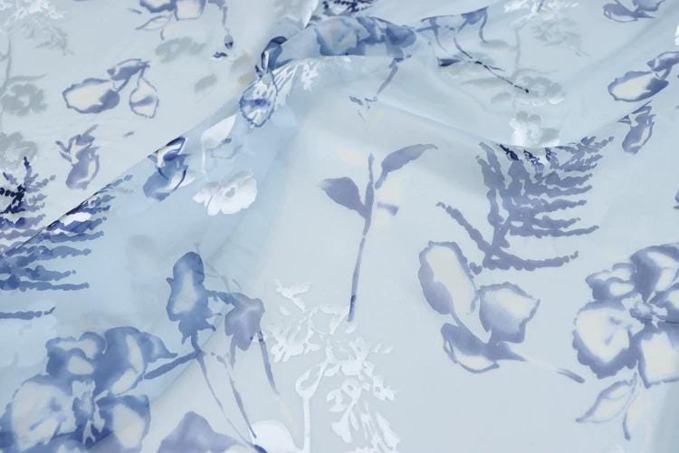 a close-up view of a fabric with a floral pattern in shades of blue and white. The fabric appears to be made of sheer material, giving it a delicate and airy appearance. The pattern consists of various flowers and leaves, creating a visually appealing and intricate design.