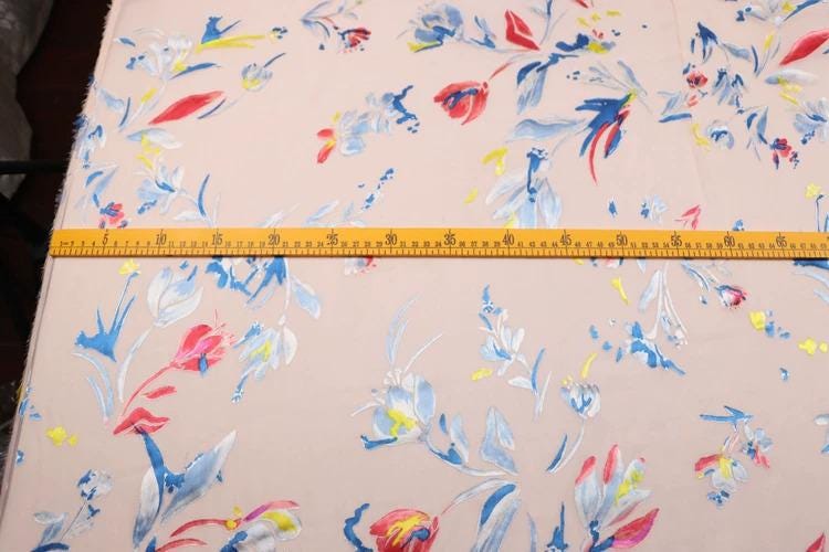 a fabric with a floral pattern, featuring various shades of pink, blue, and yellow flowers. A yellow measuring tape is placed across the fabric, likely for size reference or to measure the dimensions of the pattern.