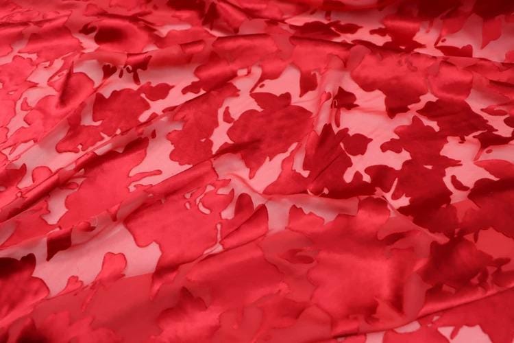 a close-up view of a red fabric with a floral pattern. The fabric appears to be made of a shiny, shiny material, possibly silk or satin, and has a textured, wrinkled appearance. The pattern consists of red flowers or leaves, which are the main visual elements of the image.