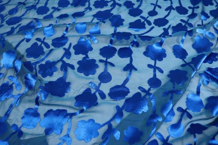 a close-up view of a fabric with a repeating pattern of blue flowers or leaves. The fabric appears to be made of a shiny, silky material, and the pattern is quite intricate and detailed.