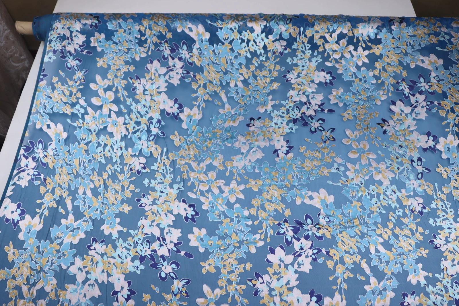 a close-up view of a fabric with a blue floral pattern. The fabric appears to be a type of silk or satin material, with a repeating pattern of blue and white flowers and leaves. The fabric is spread out on a surface, possibly a table or a countertop.