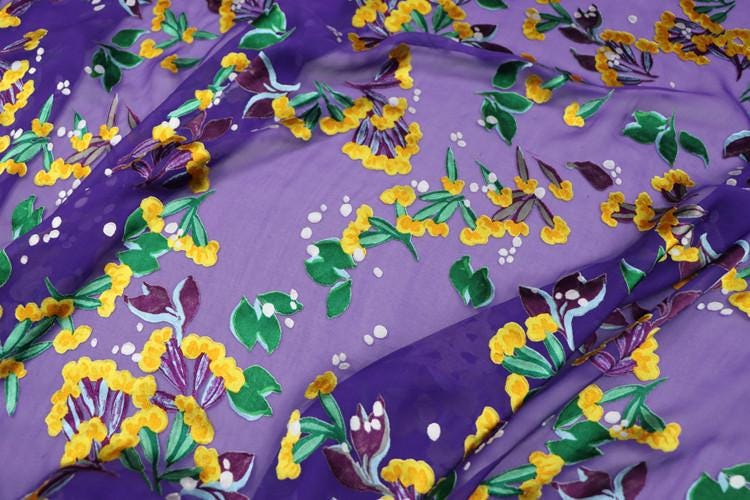 a close-up view of a fabric with a vibrant floral pattern in shades of purple and yellow. The pattern features various flowers and leaves, creating a lively and colorful design.