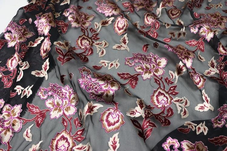 a close-up view of a fabric with a floral pattern in shades of pink, red, and gray. The fabric appears to be made of sheer material, with a black border. The pattern consists of various flowers and leaves, creating a visually appealing and intricate design.