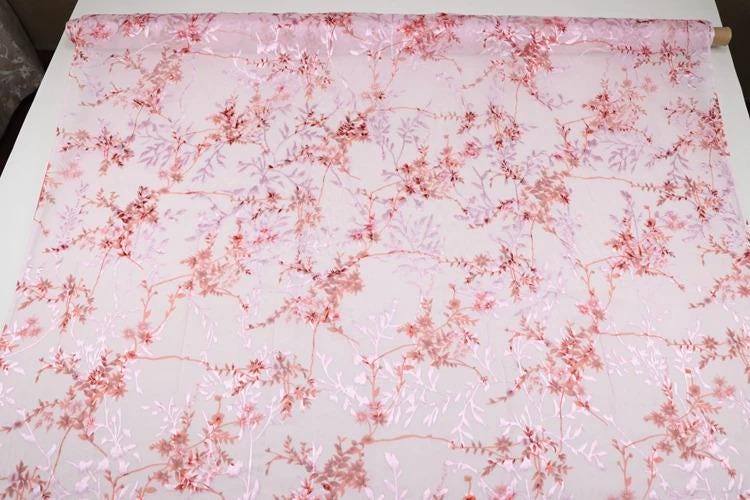 a fabric with a pink floral pattern, featuring various shades of pink and red flowers and leaves. The fabric appears to be a sheer material, with a delicate and intricate design.