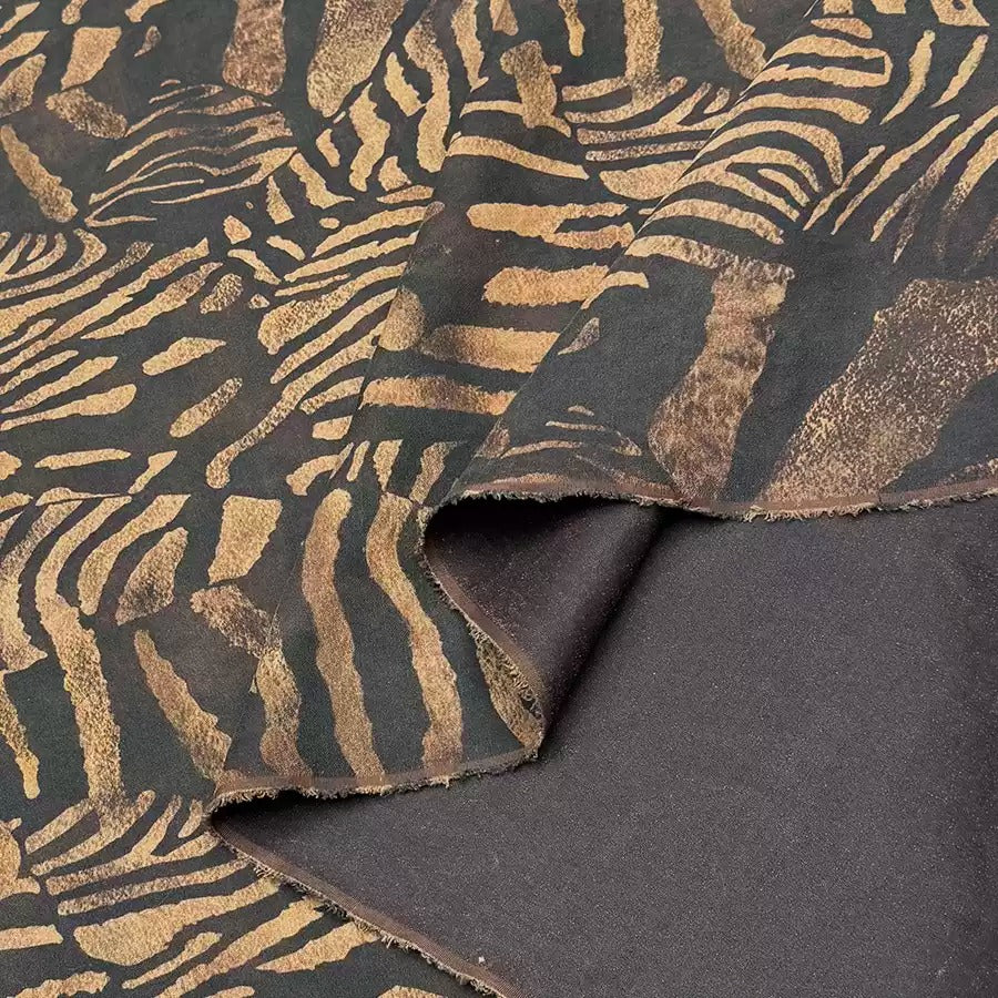 a close-up view of a fabric with a striking zebra-like pattern in shades of black and gold. The pattern is intricate and detailed, with the zebra's stripes and markings clearly visible. The fabric appears to be made of a soft, plush material, possibly cotton or a similar natural fiber. The image does not contain any text or other discernible objects, and the focus is solely on the zebra-like pattern on the fabric.