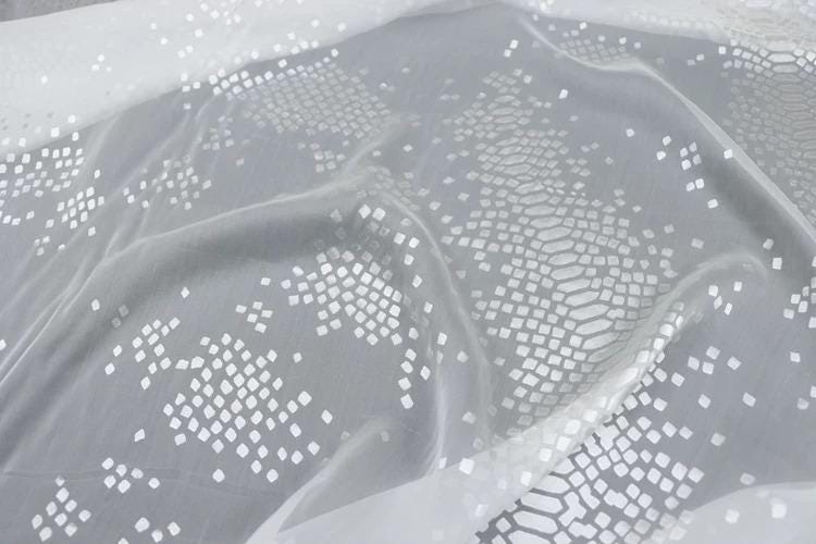 a close-up view of a fabric with a white, diamond-shaped pattern. The fabric appears to be made of a soft, flowing material, possibly silk or chiffon. The pattern consists of numerous small white diamonds arranged in a repeating geometric design, creating a visually striking and intricate appearance.