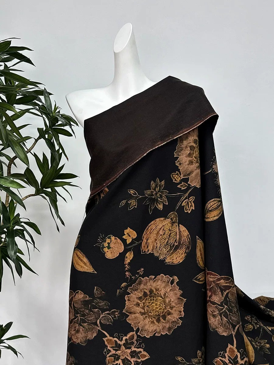 A mannequin wearing a black and gold floral print dress with a brown shawl draped over it, placed next to a potted plant.
