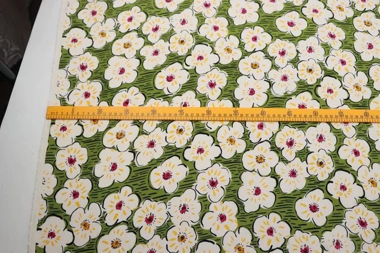 a fabric with a floral pattern, featuring white and green flowers with yellow centers. A yellow ruler is placed across the fabric, indicating the size or dimensions of the pattern.
