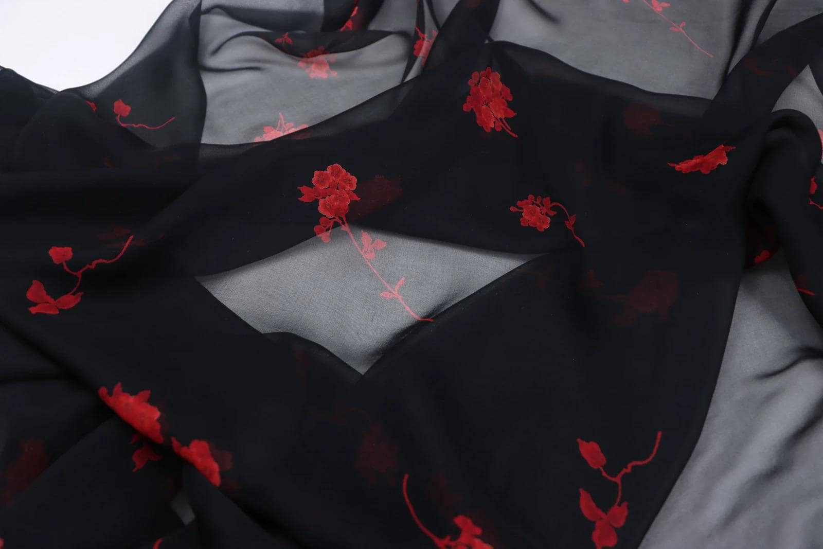 a close-up view of a black fabric with red floral embroidered patterns. The fabric appears to be made of sheer material, giving it a delicate and flowing appearance. The red flowers are scattered across the fabric, creating a visually striking contrast against the black background.