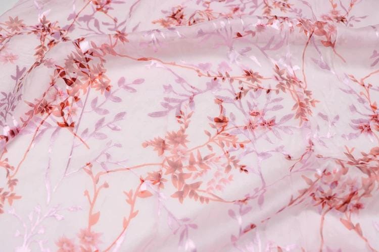 a close-up view of a fabric with a pink floral pattern. The fabric appears to be made of sheer material, with delicate pink flowers and leaves covering the entire surface. The pattern is intricate and detailed, creating a visually appealing and textured appearance.