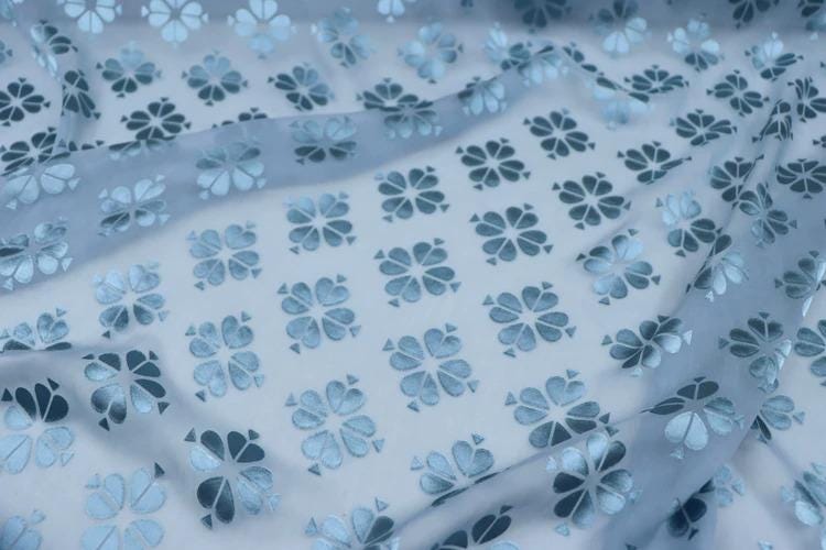 a close-up view of a fabric with a repeating pattern of blue and white flowers or leaves. The pattern is intricate and detailed, with each flower or leaf being a small, symmetrical design. The fabric appears to be made of a soft, flowing material, possibly silk or chiffon, and has a slightly wrinkled texture. The pattern repeats itself across the entire fabric, creating a visually striking and harmonious design.