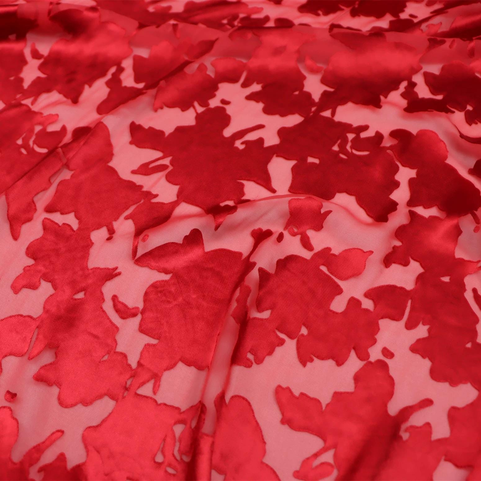 a close-up view of a fabric with a red floral pattern. The fabric appears to be made of a shiny, satin-like material, and the pattern consists of red flowers or leaves against a pink background. The fabric is folded and crumpled, giving it a textured appearance.