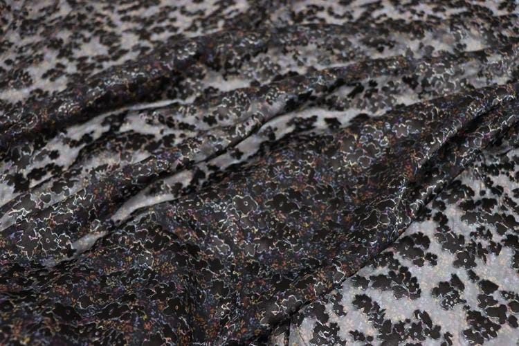 a close-up view of a fabric with a black and white floral pattern. The fabric appears to be made of a shiny material, possibly silk or satin, and has a textured appearance. The pattern consists of small black and white flowers or leaves, creating a visually striking contrast against the dark background.