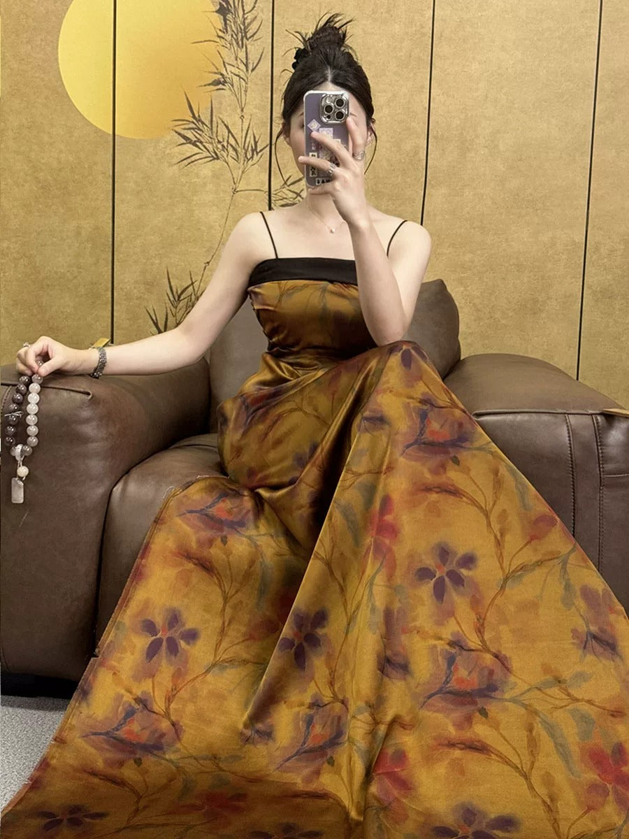 A woman in a long, floral-patterned dress is sitting on a couch, holding a phone and taking a selfie.