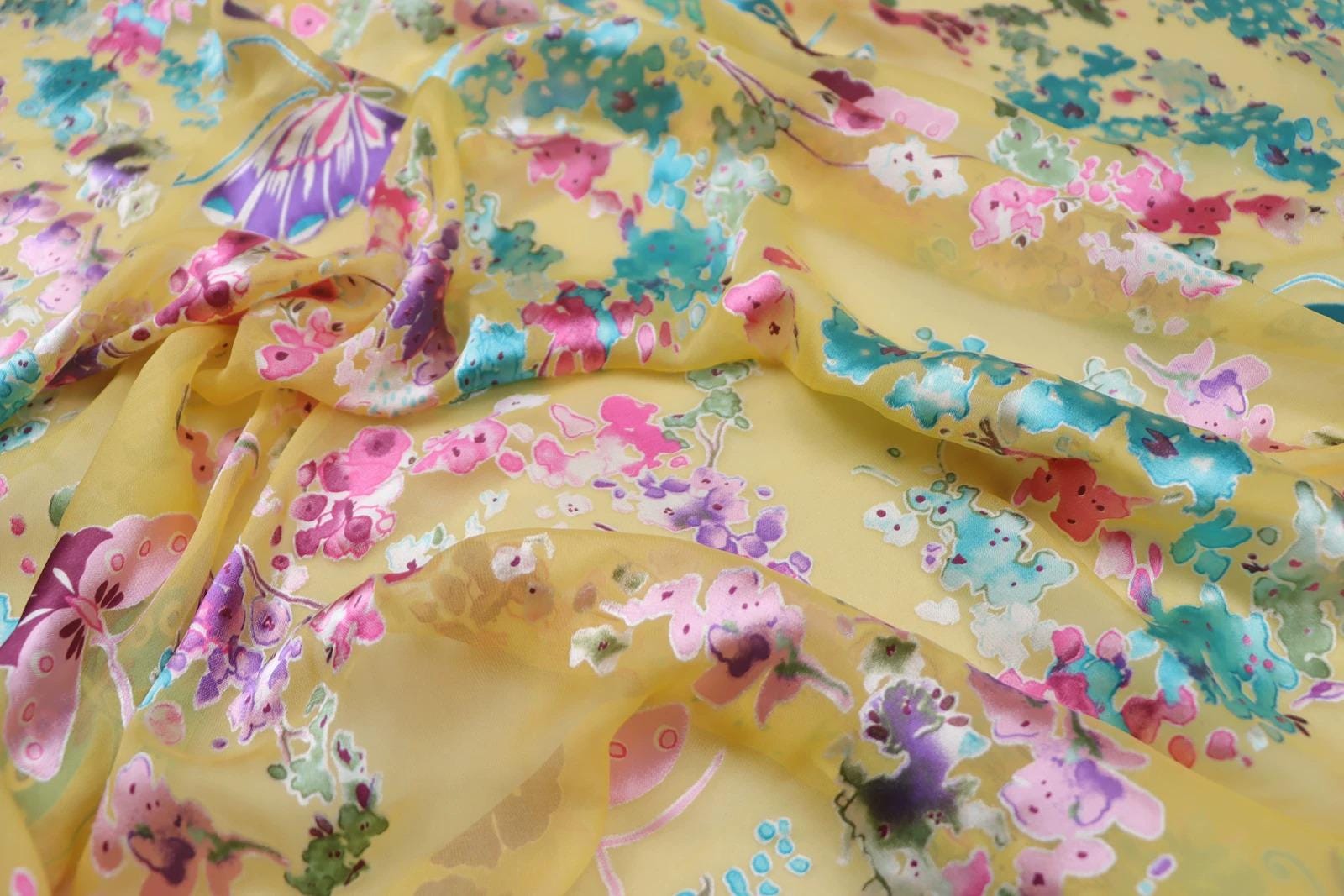 a close-up view of a fabric with a vibrant, colorful floral pattern. The fabric appears to be made of silk or chiffon, and the pattern consists of various flowers and leaves in shades of pink, blue, green, and yellow. The fabric is crumpled and wrinkled, giving it a soft and delicate appearance.