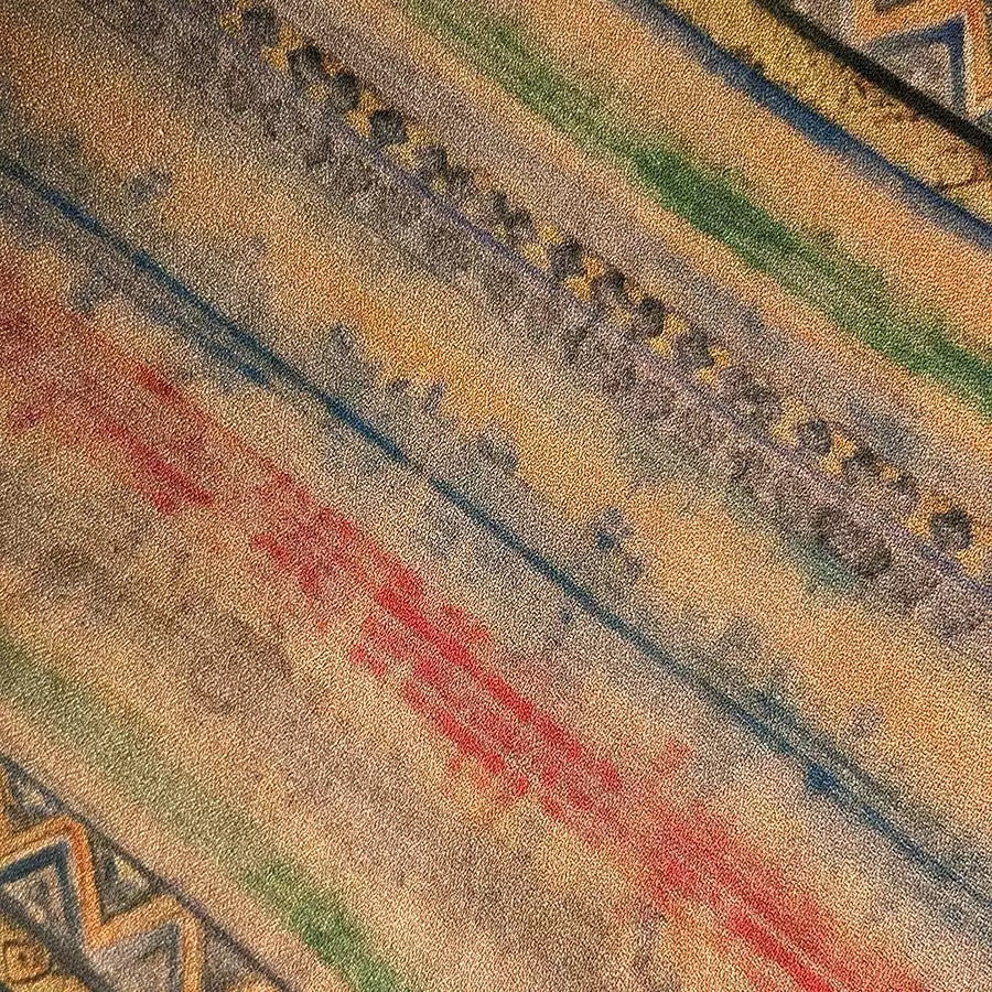 The image is a close-up of a colorful, abstract pattern with various shades of red, blue, green, and yellow. The pattern appears to be a textile or fabric design, with a diagonal arrangement of the different colors creating a visually striking and dynamic composition.