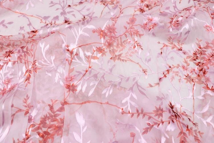 a close-up view of a fabric with a pink floral pattern. The fabric appears to be made of sheer material, with delicate pink flowers and leaves covering the entire surface. The pattern is intricate and detailed, showcasing the craftsmanship of the fabric.