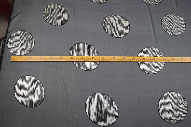 a gray fabric with a repeating pattern of black and white circles, and a yellow measuring tape is laid across the fabric, indicating the size or dimensions of the pattern.