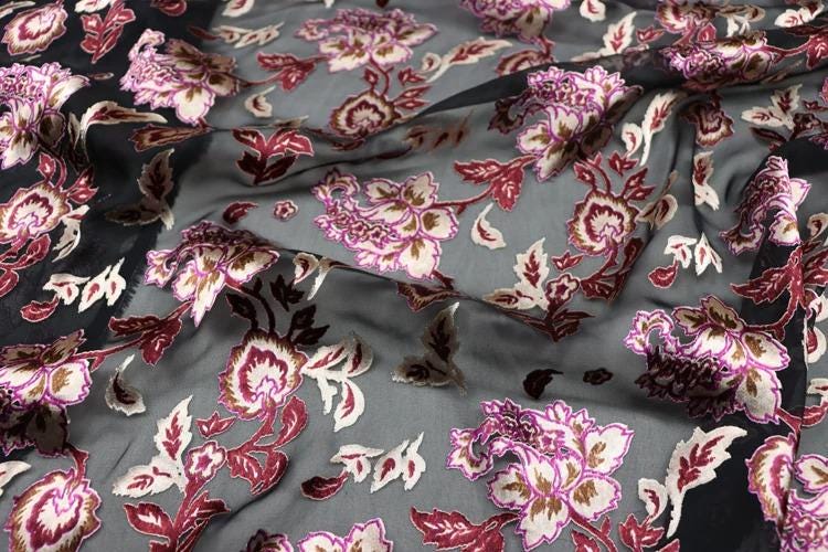 a close-up view of a fabric with a floral pattern in shades of pink and red. The fabric appears to be made of silk or chiffon, and the pattern consists of various flowers and leaves in different sizes and colors. The fabric is crumpled, giving it a textured appearance.