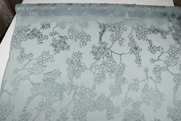 a close-up view of a fabric with a repeating pattern of green floral designs. The pattern consists of various shades of green, with some areas appearing darker and others lighter, creating a visually interesting and intricate design.
