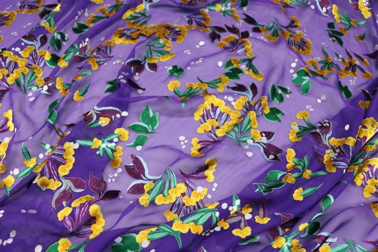 a close-up view of a fabric with a vibrant floral pattern in shades of purple, yellow, and green. The pattern is composed of various flowers and leaves, creating a visually striking and colorful design.