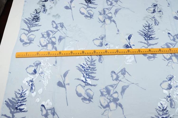 a fabric with a blue and white floral pattern, and a yellow measuring tape is placed across it, indicating the size of the fabric.
