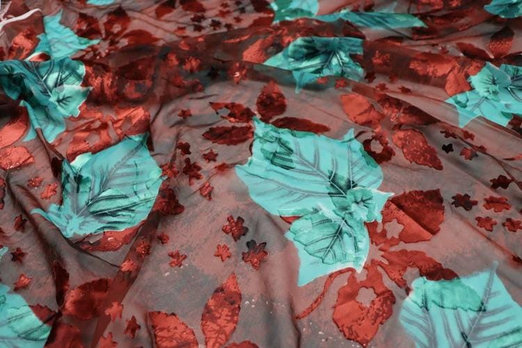 a close-up view of a fabric with a floral pattern. The fabric has a mix of red and green colors, with some areas appearing to be red and others green. The pattern consists of various shapes and sizes of leaves and flowers, creating a visually interesting and textured appearance.